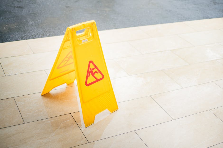 A bright yellow, freestanding wet floor caution sign with red warning symbols is placed on a beige tiled floor near a corner. The sign displays an icon of a person slipping, indicating a potentially s
