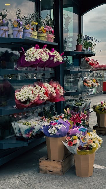A display of flower bouquets on black metal shelving outside a florist shop, with arrangements featuring white, pink, purple, and yellow flowers wrapped in colorful paper, some in brown kraft paper, a