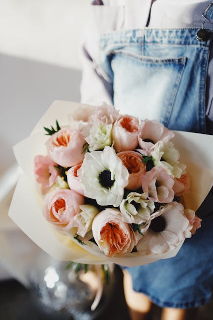 A person wearing a white top and blue denim jeans holds a bouquet of fresh flowers wrapped in cream-colored paper. The arrangement features soft pink roses, white anemones with dark centers, and peach