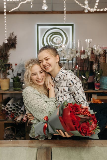 Two young women are smiling and embracing each other in a flower workshop environment, with one holding a large, elaborate floral arrangement wrapped in grey and red paper, featuring vibrant red flowe