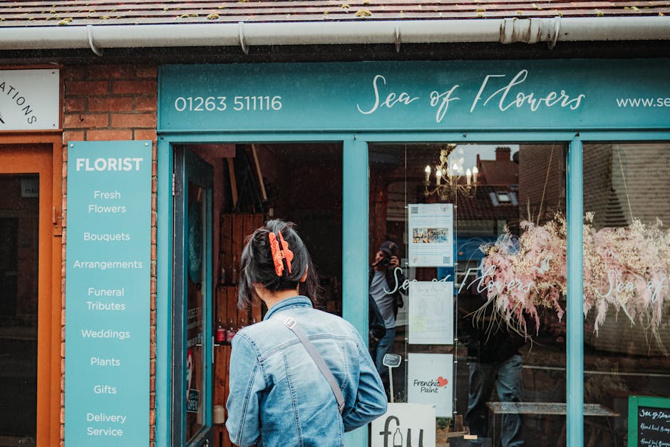 A woman with short dark hair secured by an orange hair clip stands outside the storefront of 'Sea of Flowers', a flower shop with a teal-colored awning displaying the shop's name and contact number. T
