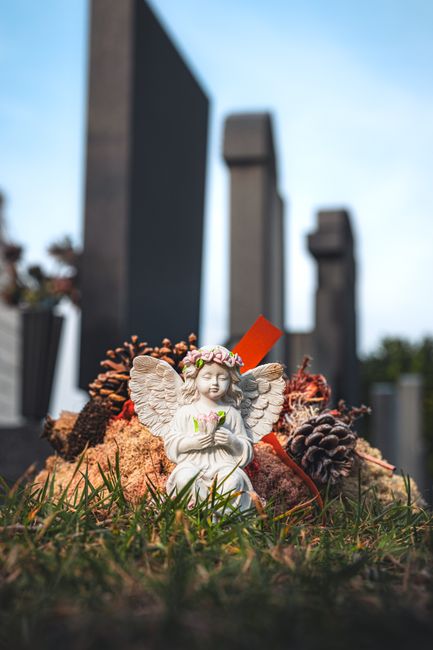 A ceramic angel figurine with detailed wings, wearing a floral wreath crown and holding a small bouquet of pink and green flowers, is placed among pinecones, dried leaves, and a reddish-orange ribbon 
