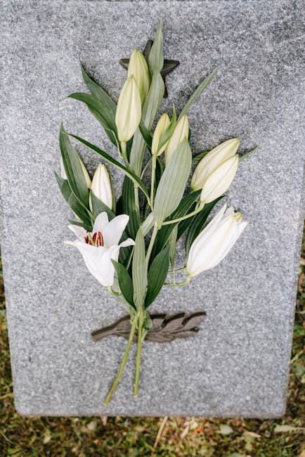 A delicate floral arrangement of white lilies and lilies of the valley with elongated green leaves, affixed to a grey granite memorial stone. The lilies are in full bloom, showcasing their smooth, tru