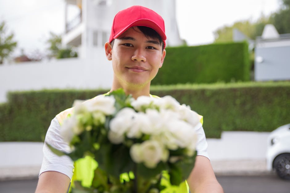A young man wearing a red cap and a white t-shirt, smiling and holding a bouquet of fresh white flowers with green leaves, presented in a simple wrapping. The bouquet features a mix of petal textures