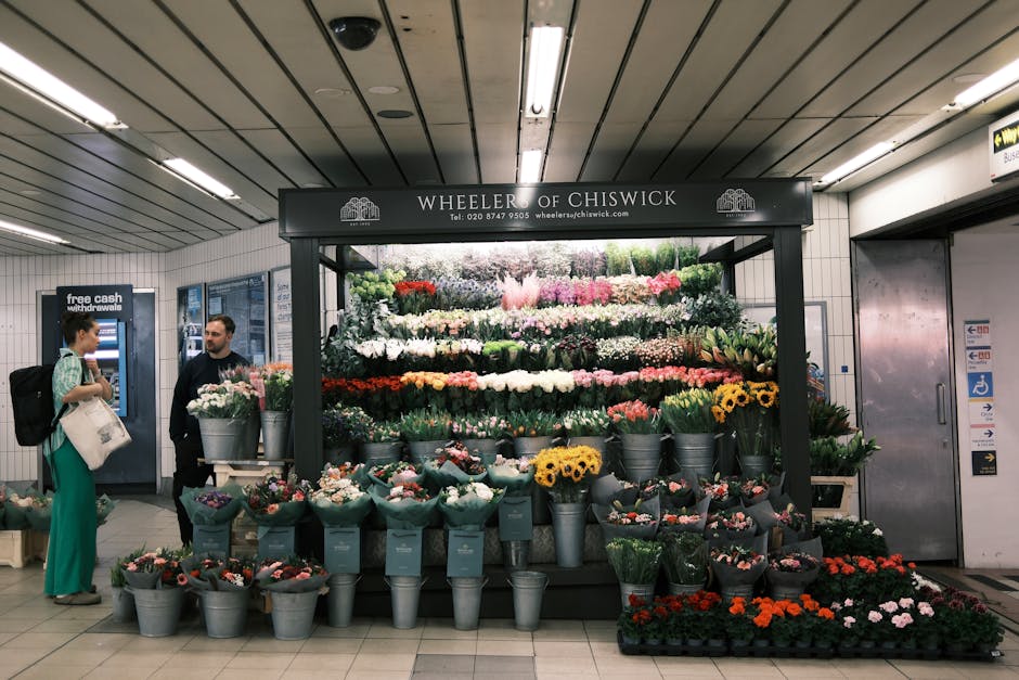A flower stall labeled 'Wheeler of Chiswick' located in a subway station, displaying a vibrant and varied arrangement of fresh flowers. The arrangement includes roses, sunflowers, tulips, and other se