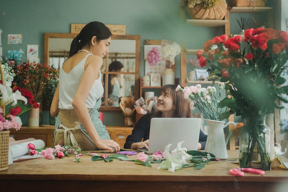 A woman in a white sleeveless top and beige apron stands at a wooden table, engaging in conversation with a smiling woman seated behind a laptop. The seated woman has shoulder-length brown hair and is