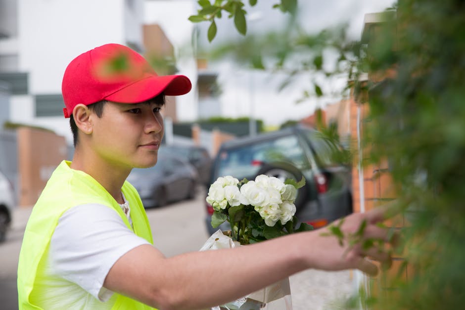 A young male florist wearing a red cap and a high-visibility yellow vest is seen holding a bouquet of white roses with lush green stems and leaves. He is placing the floral arrangement against a leafy